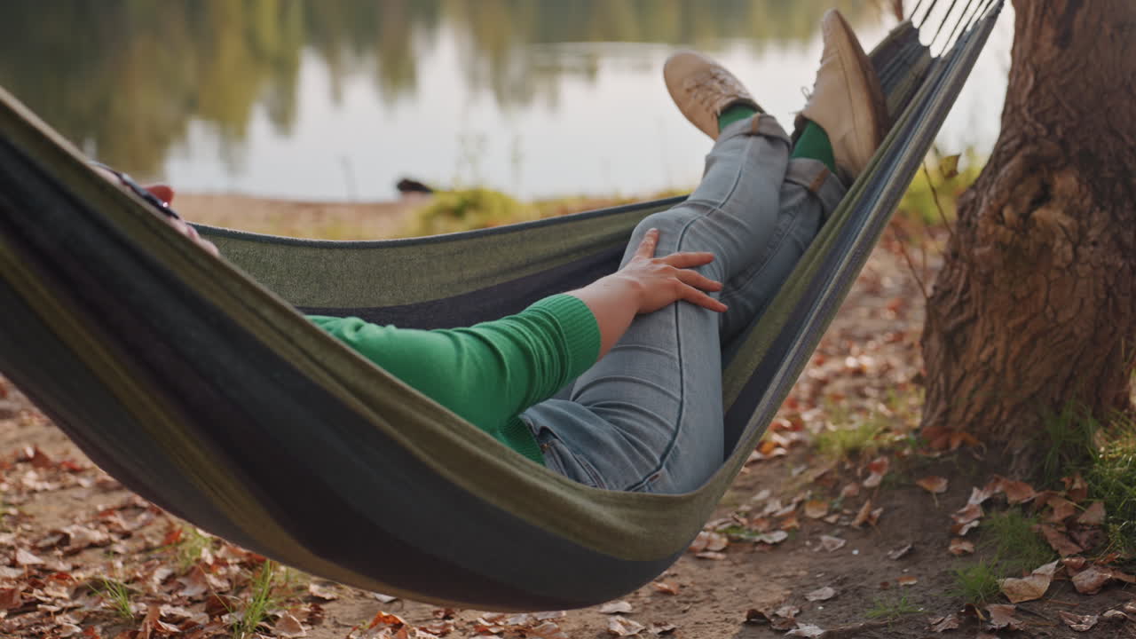 Donna bianca che sonnecchia sull'amaca in riva al lago, maglione verde, occhiali da sole, jeans, tronco d'albero come ancoraggio accanto all'acqua, lievi increspature, luce dorata, atmosfera accogliente di pisolino in campeggio, fotografo in pausa per un riposo creativo