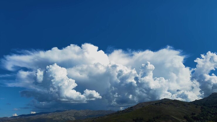 huge, billowing white cumulus clouds accumulating rapidly over a rolling mountain peak under a bright blue sky.