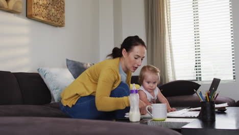 Madre-Caucásica-Sosteniendo-A-Su-Bebé-Usando-Una-Computadora-Portátil-Y-Tomando-Notas-Mientras-Trabaja-Desde-Casa