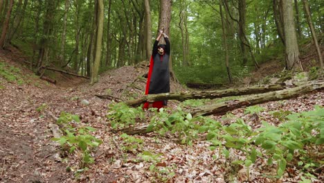 Parca-Con-Cráneo-Animal-Con-Capa-Negra-Y-Roja-Encapuchada,-Parada-En-El-Bosque-Y-Golpeando-La-Guadaña-En-El-Suelo