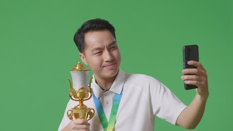 골드 트로피를 들고 스마트폰으로 사진을 찍는 금메달을  아시아 남자가 스튜디오에서 초록색 화면 배경에서 첫 번째 우승자로 화면을 찍습니다.