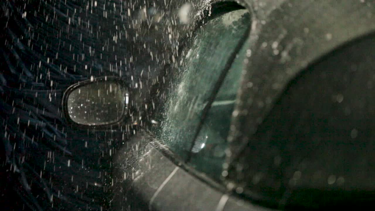 From the interior perspective, a handheld camera focuses on a car's rectangular side mirror, in a bustling car wash framed by blue bristles and dancing water droplets