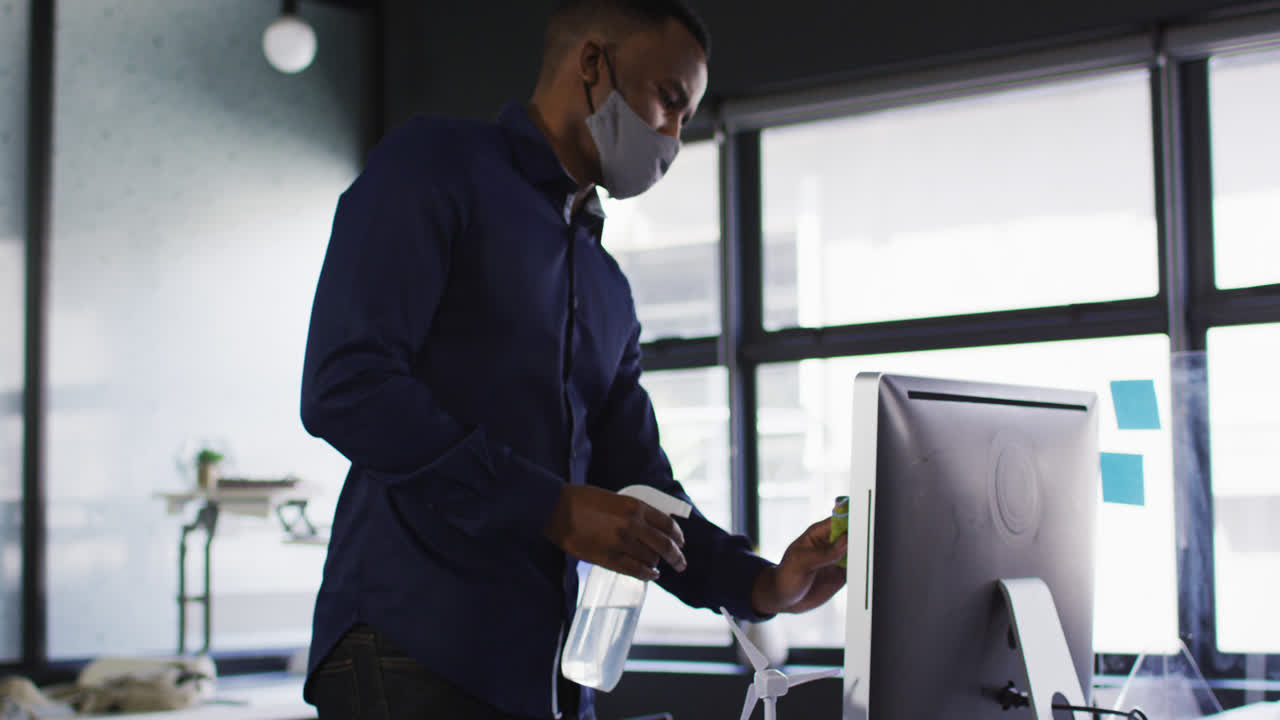 African american man wearing face mask cleaning his computer with disinfectant at modern office