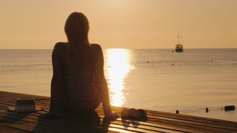 An-Adult-Woman-Sits-In-A-Summer-Sarafan-On-The-Beach-Sits-On-A-Bench-And-Holds-Onto-A-Suitcase-On-Wh