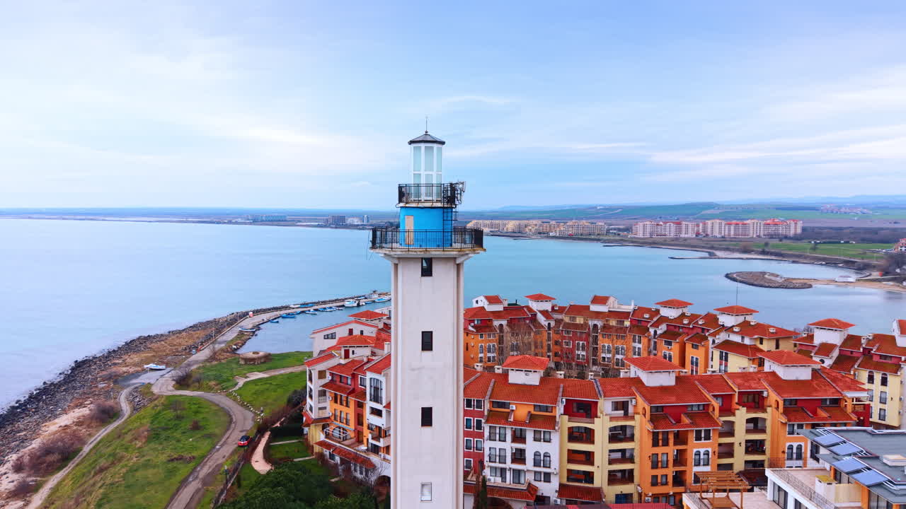 Faro contro lo sfondo di una città costiera. Inquadratura aerea media di un faro bianco con edifici e il mare sullo sfondo sotto un cielo nuvoloso.