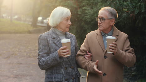 elderly married couple walking in the park, talking and drinking hot coffee at sunset in autumn