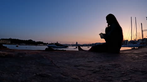 mujer sentada junto a un puerto con la puesta de sol en el fondo