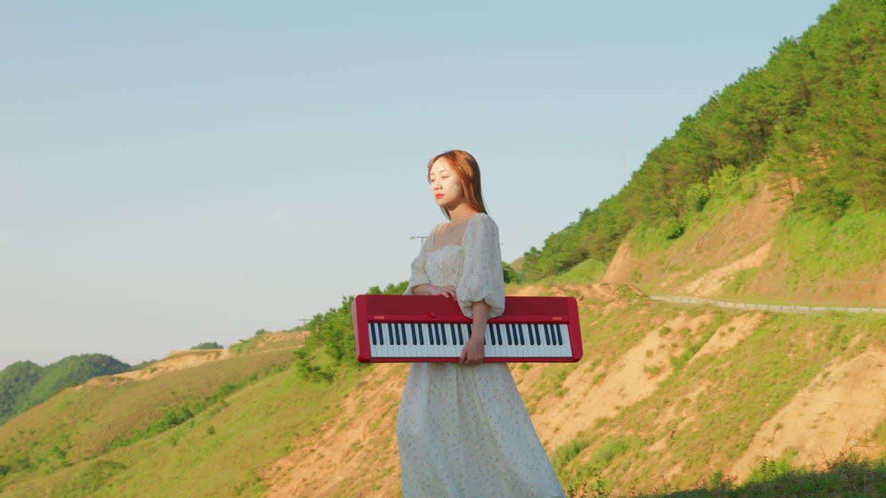 young musician artist carrying a red piano keyboard musical instrument in nature on mountain path wearing long white elegant dress