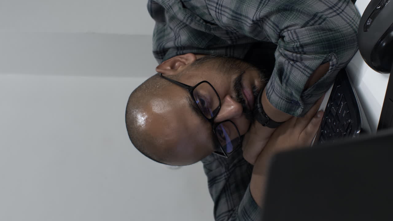 Vertical close up shot of an exhausted overworked young Indian entrepreneur as he rests his head on his arms in front of a computer, his eyes fixed on the screen waiting for an important email