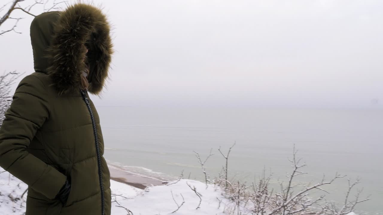 Young woman in green winter clothes standing on the steep dunes along the sandy shore of the Baltic sea beach, old military building ruins, medium tracking shot