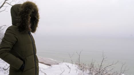 young woman in green winter clothes standing on the steep dunes along the sandy shore of the baltic sea beach, old military building ruins, medium tracking shot