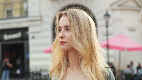 Retrato-De-La-Encantadora-Joven-Rubia-Mirando-A-Un-Lado-Y-Girando-La-Cara-Hacia-La-Cámara-Con-Una-Sonrisa-En-El-Centro-De-La-Ciudad