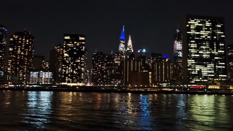 panoramic view to the left of new york city at night from a river.
