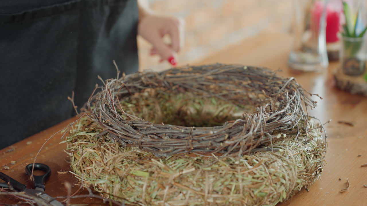 Female florist arranging handmade wreath using twigs and hay on wooden table with scissors and natural materials, demonstrating detailed process of rustic decoration crafting with focus
