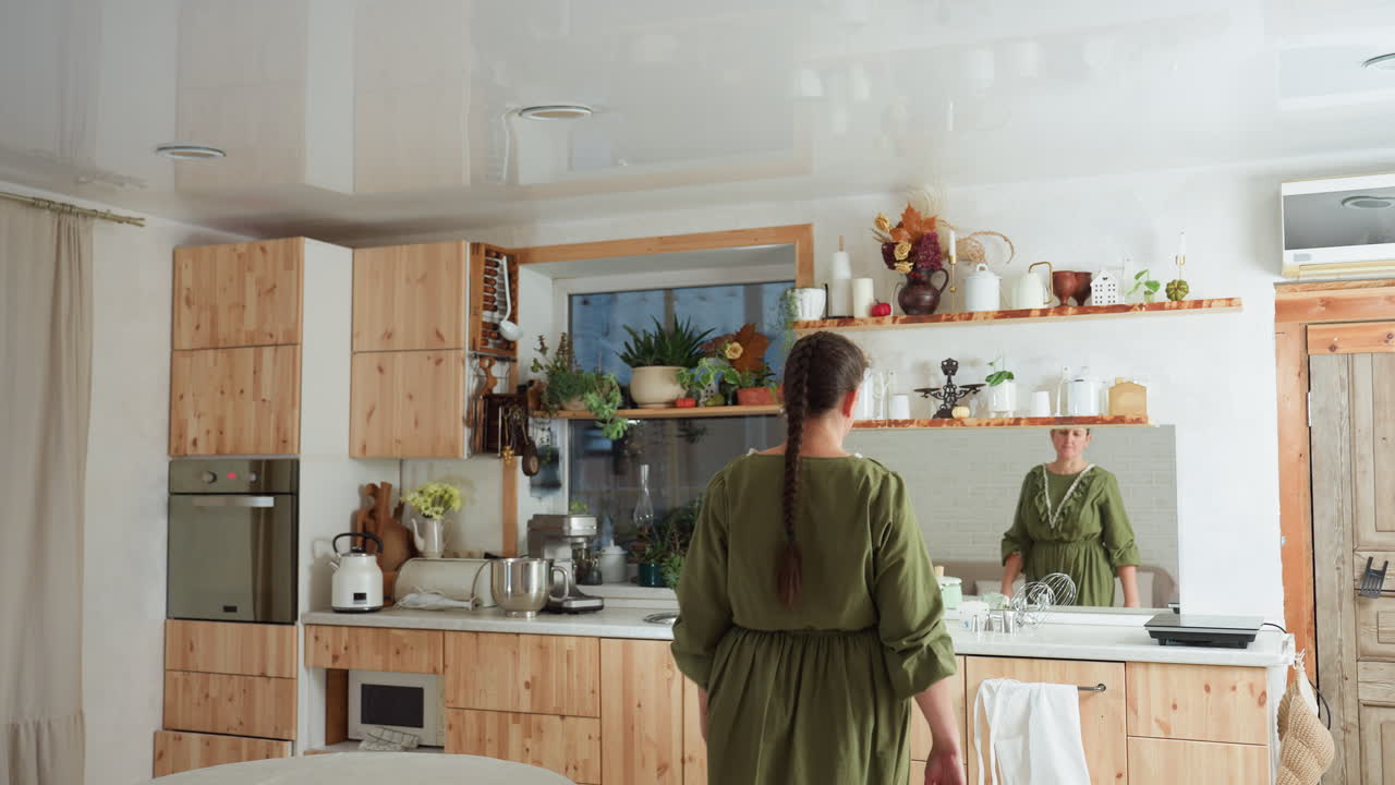 Woman with braided hair dressed in green enters rustic kitchen filled with indoor plants, wooden shelves, and decor while her reflection is visible in mirror beside white sink and wall-mounted items