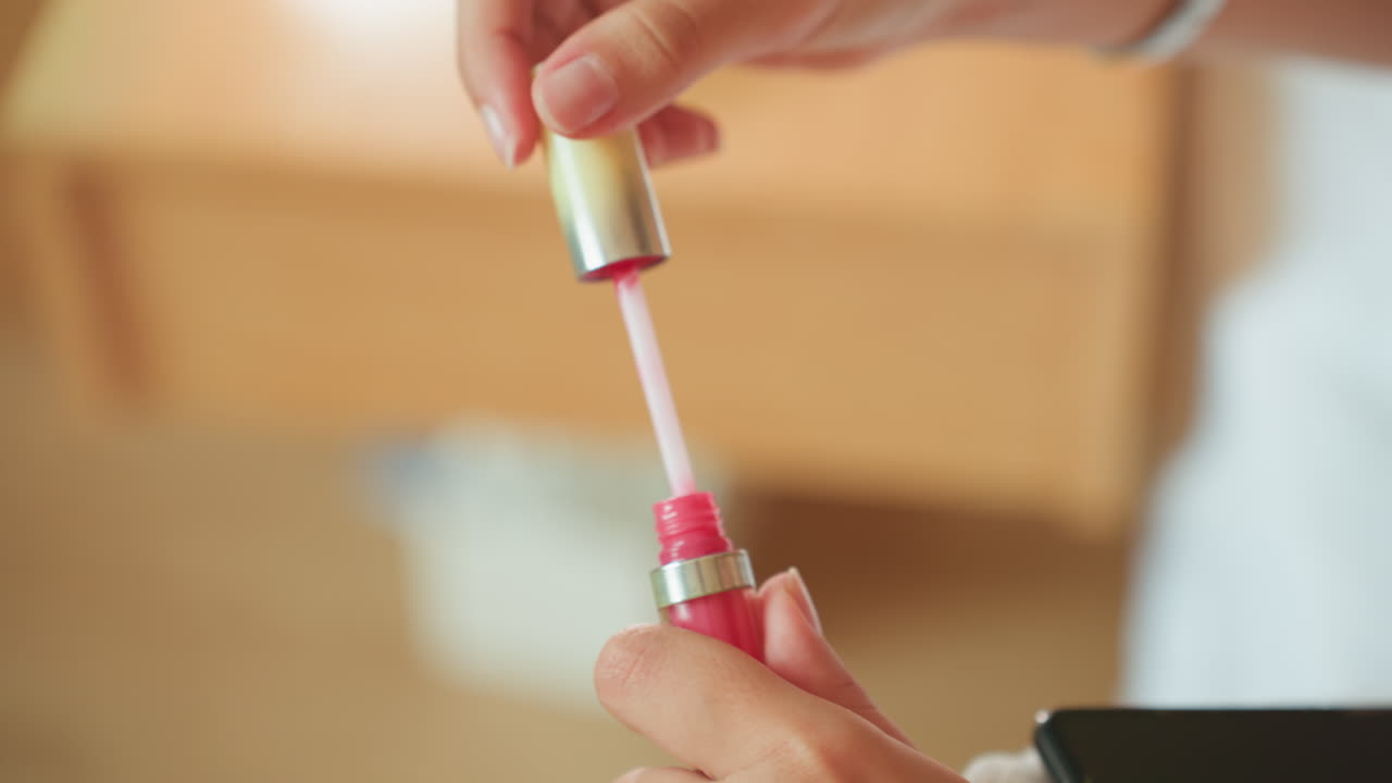 Gæst åbner lyserød lipgloss på hotelværelse, nærbillede af hænder, der fjerner hætten og løfter applikatoren, skinnende pigment på staven, blødt morgenlys, sløret kommodebaggrund, intim rejse-skønhedsrutine