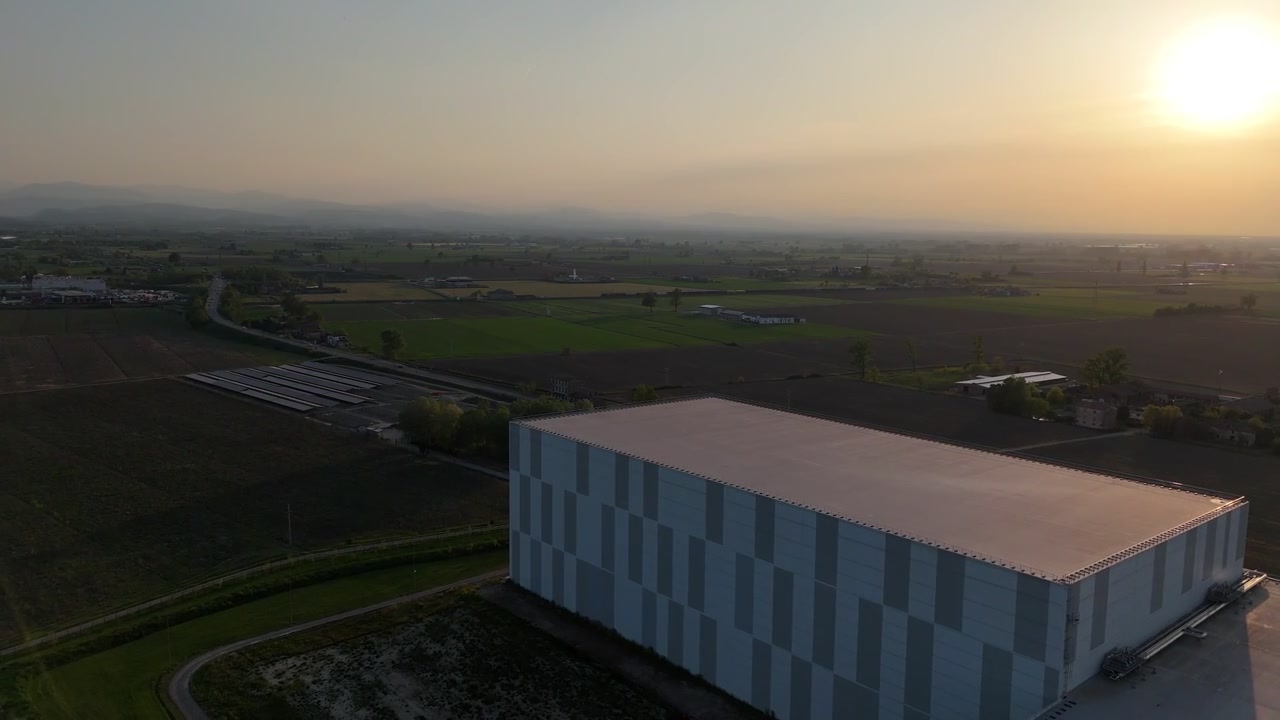Automatiseret kølelager med isoleret facade ved siden af landbrugsmarker og solcelleanlæg på Po-sletten i solnedgangens lys, der viser logistikinfrastruktur og temperaturforhold.