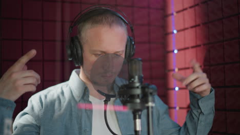 a man wearing a blue long sleeve shirt, white undershirt, and black headphones is standing in front of a microphone, making hand movements in a red soundproofed recording room