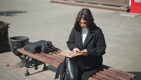 young woman sits outdoors on a bench reading a book with yellow-edged pages, she flips through the pages thoughtfully while enjoying the fresh air and sunlight