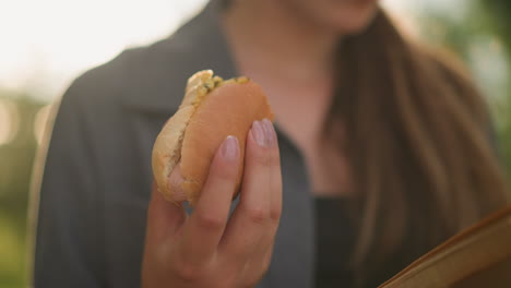 primer plano de una mujer con una camiseta gris sosteniendo un shawarma mientras lee un libro en un entorno al aire libre iluminado por el sol, la luz del sol suave ilumina su mano y el bocadillo, con un fondo borroso de vegetación y cálidos rayos de sol