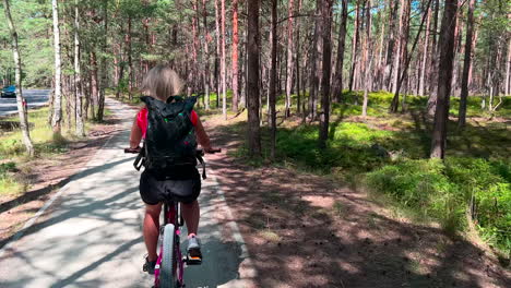 Mujer-Adulta-Joven-En-Bicicleta-Por-El-Parque-De-Pinos-De-La-Ciudad---Seguimiento-De-La-Vista-Trasera