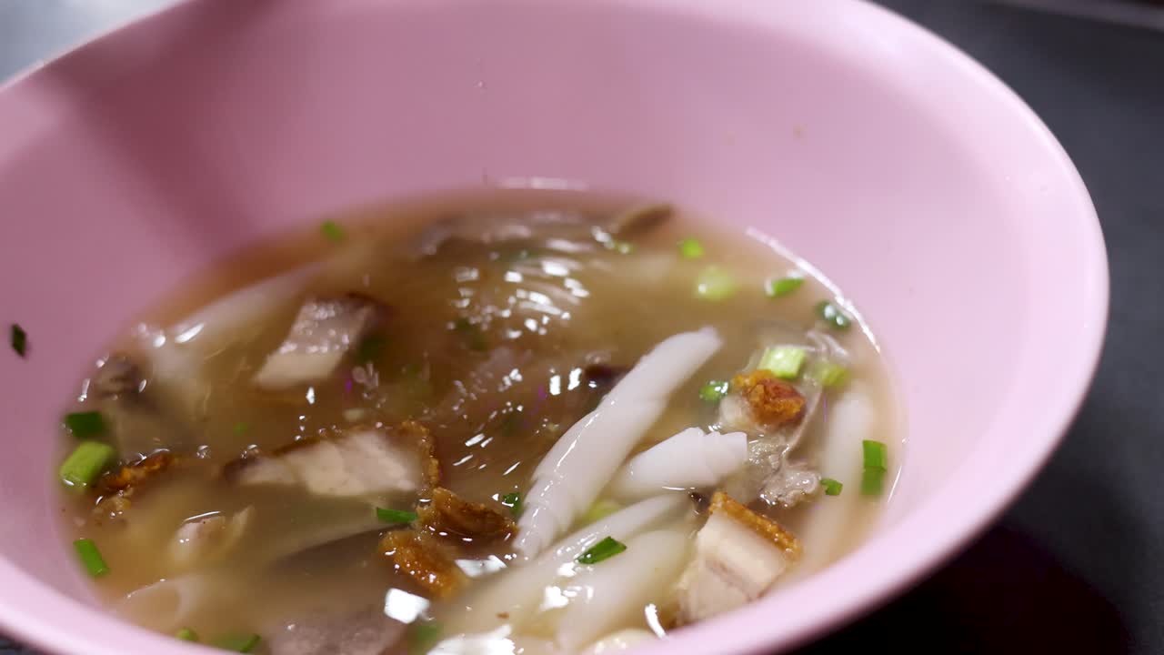 Hand stirs rolled rice noodle soup with crispy pork in pink bowl, close-up, street food