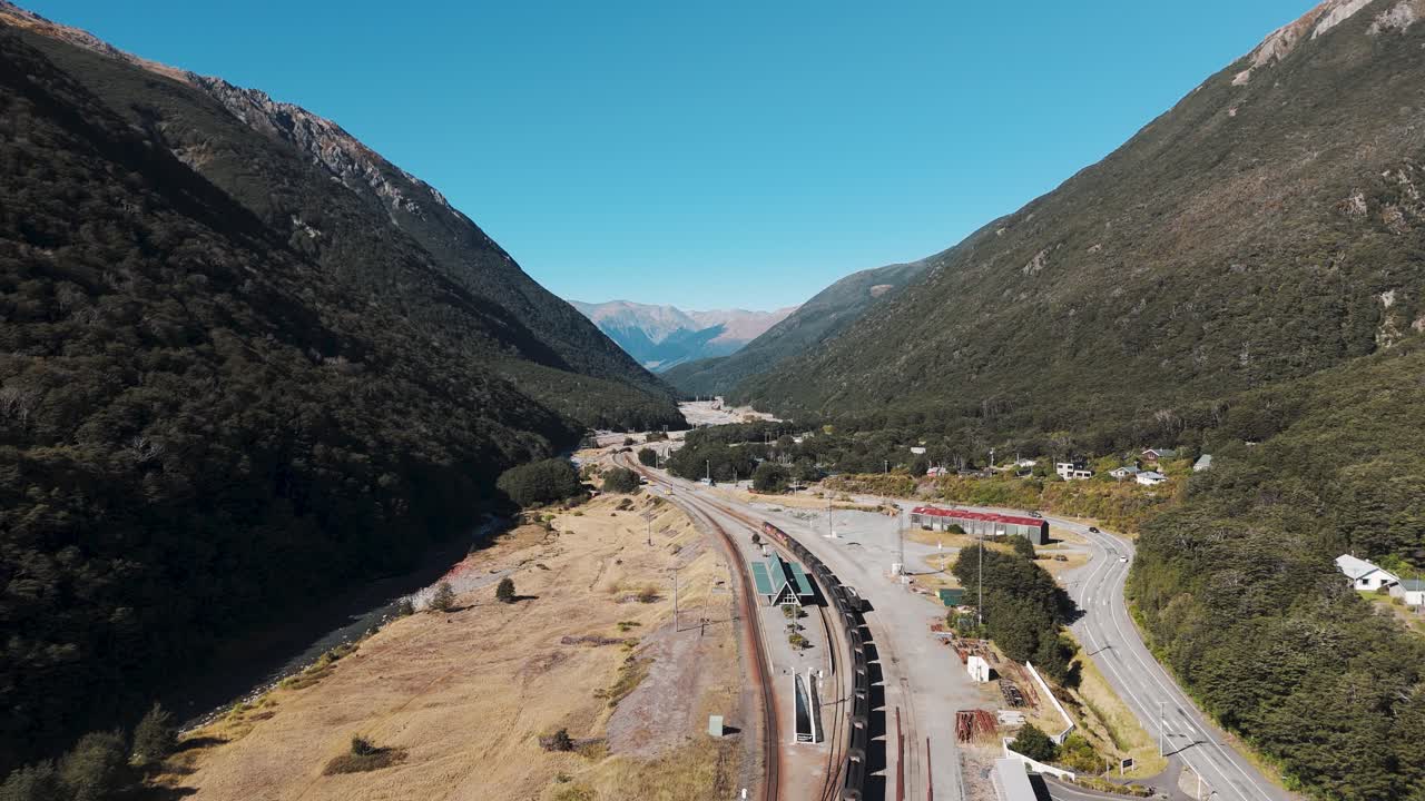 Arthur’s Pass Train Station, New Zealand’s Highest Elevation Stop. Aerial backwards weide shot. Mountains and blue sky in summer. Cars on highway.
