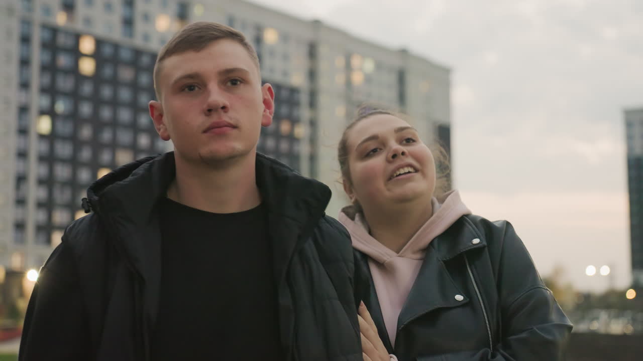 Sober boyfriend walks quietly with cheerful girlfriend who gently rests her head on his shoulder showing comfort and connection during evening stroll with tall buildings in soft evening background