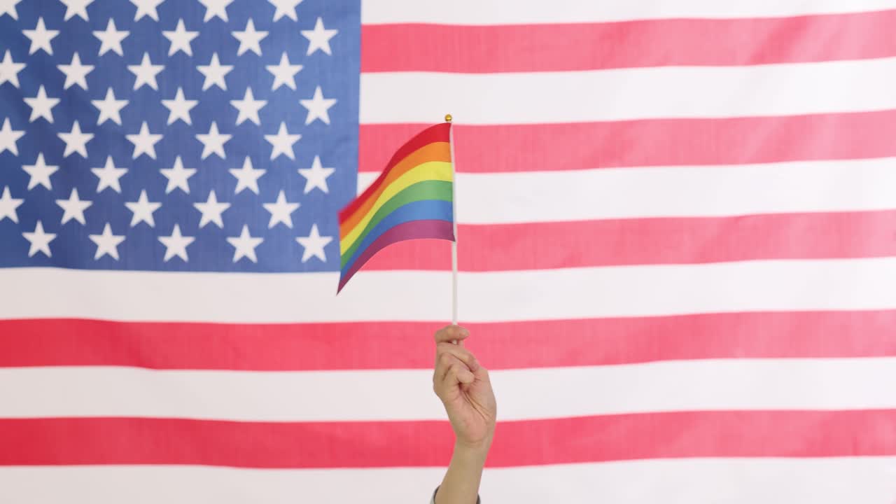Hand holding a rainbow flag in front of an American flag