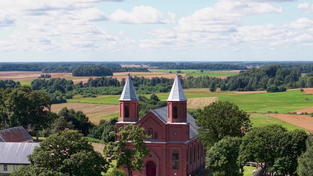 Fantastiska flygbilder visar en röd tegelkyrka med två spiror, omgiven av frodiga gröna fält och träd under en klarblå himmel, fångad i livfullt dagsljus och med skarpa, stabila detaljer.