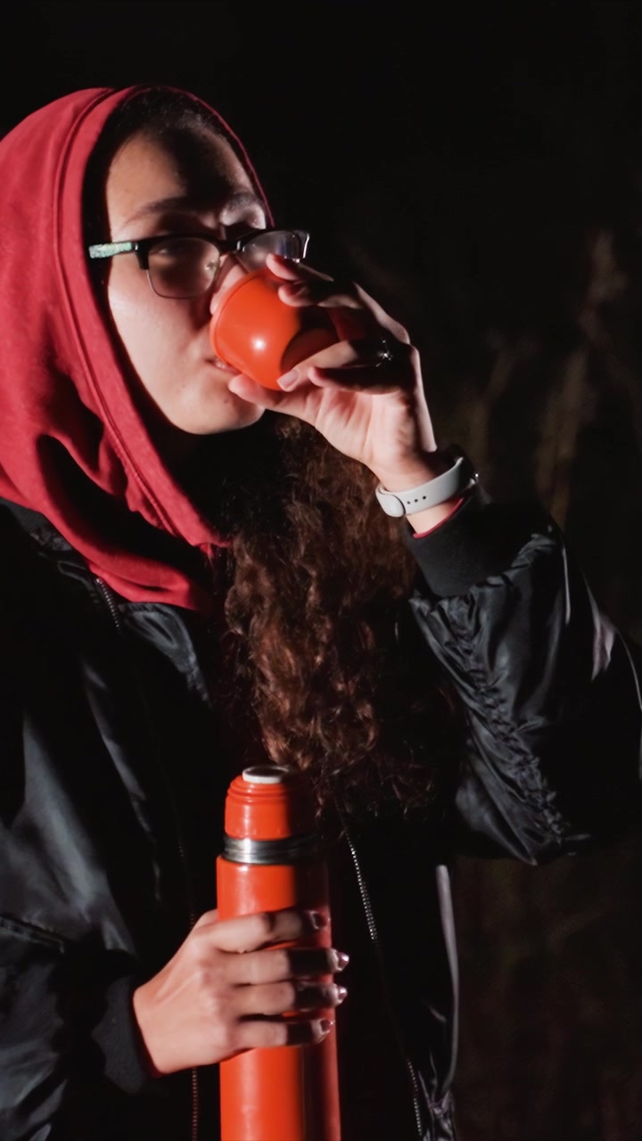 Person in dark clothing grasps both beverage containers. Orange thermos features silver band while cup emits. Dark background suggests outdoor night setting or indoor low