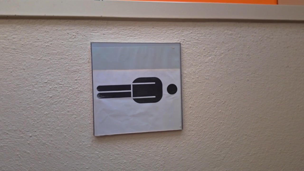 Turning sign on textured wall showing a stylized horizontal male restroom icon, black on white, slightly tilted