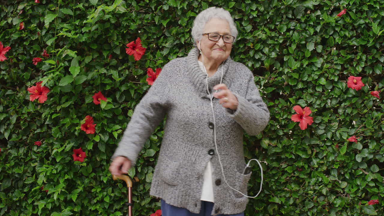 portrait of lively elderly woman dancing wearing earphones listening to music laughing enjoying playful fun in beautiful green garden flower wall outdoors