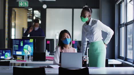 african american woman and asian woman wearing face masks discussing over laptop at modern office