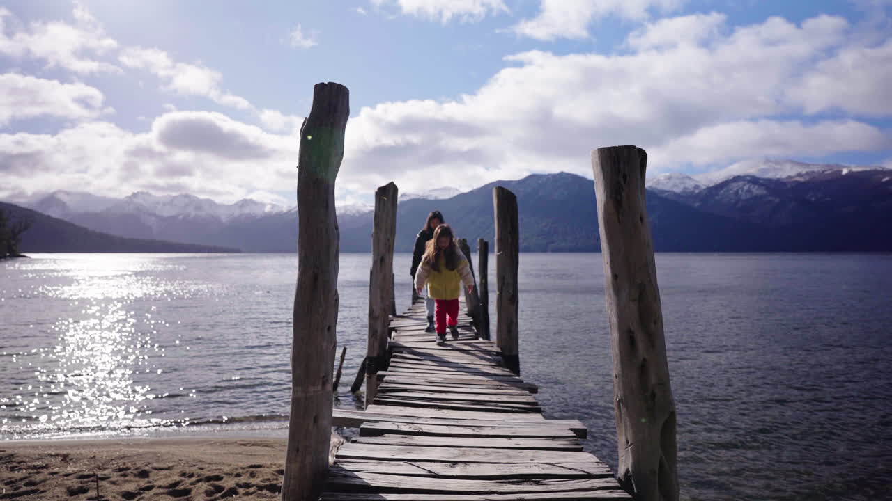 Mother and daughter walk on a rustic pier, with misty mountains and calm water below a sunny, cloud-streaked sky