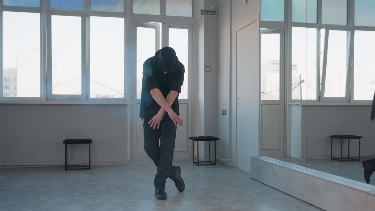 young man in black shirt and vest dances elegantly in spacious studio with rising smoke from fog machine behind him casting moody atmospheric haze across reflective floor and glass wall