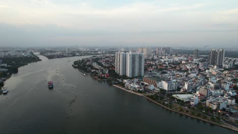 Luftaufnahme-Eines-Frachtschiffs,-Das-Auf-Dem-Saigon-Fluss-Fährt,-Mit-Hohen-Gebäuden-Im-Hintergrund-Bei-Sonnenuntergang-In-Vietnam