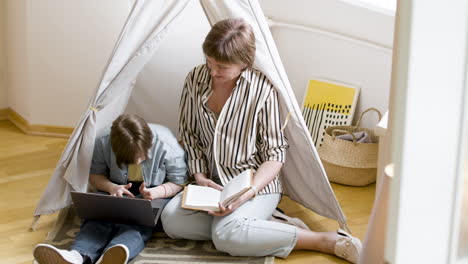 mother reading book at home next to her daughter using laptop