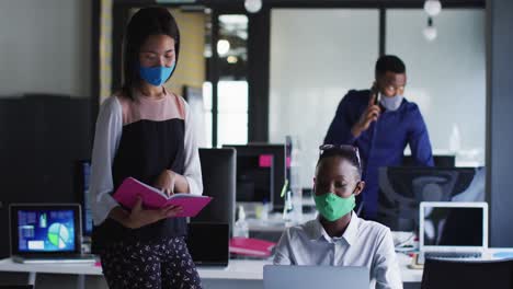 African-american-woman-and-asian-woman-wearing-face-masks-discussing-over-book-at-modern-office
