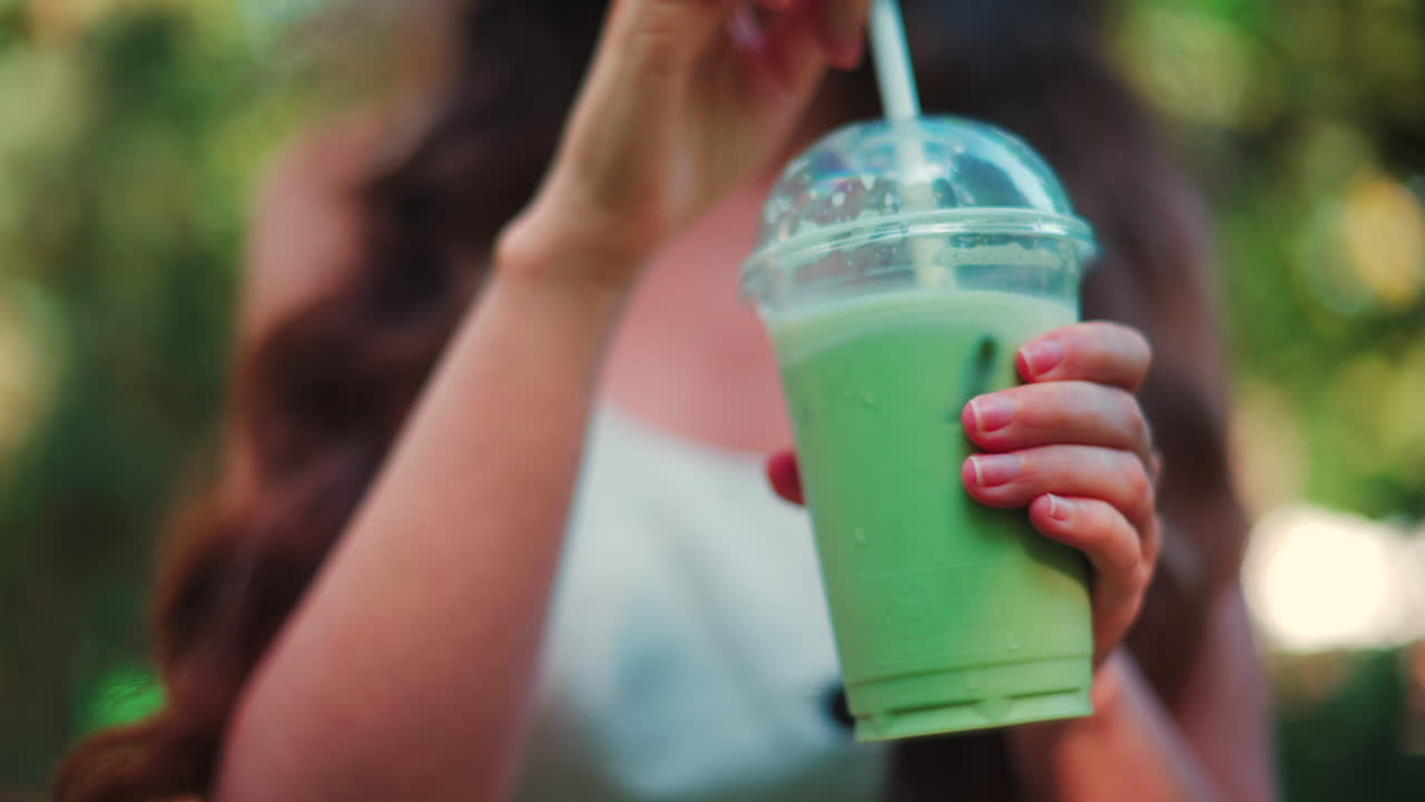 Close up of a woman drinking an iced matcha in the park, in sunlight