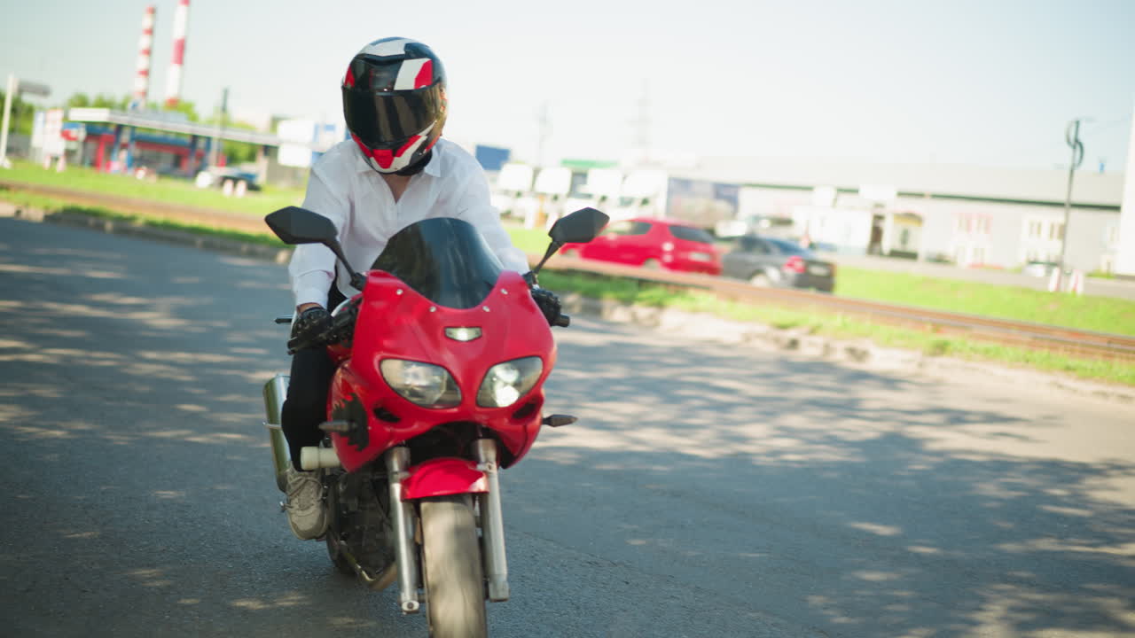 una mujer motociclista monta una bicicleta eléctrica roja, haciendo una curva en una carretera sombreada con un coche siguiendo detrás, otros coches son visibles a través de la carretera con edificios vistos a través del camino