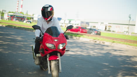 a female motorcyclist rides a red power bike, making a turn on a shaded road with a car following behind, other cars are visible across the road with buildings seen across the road