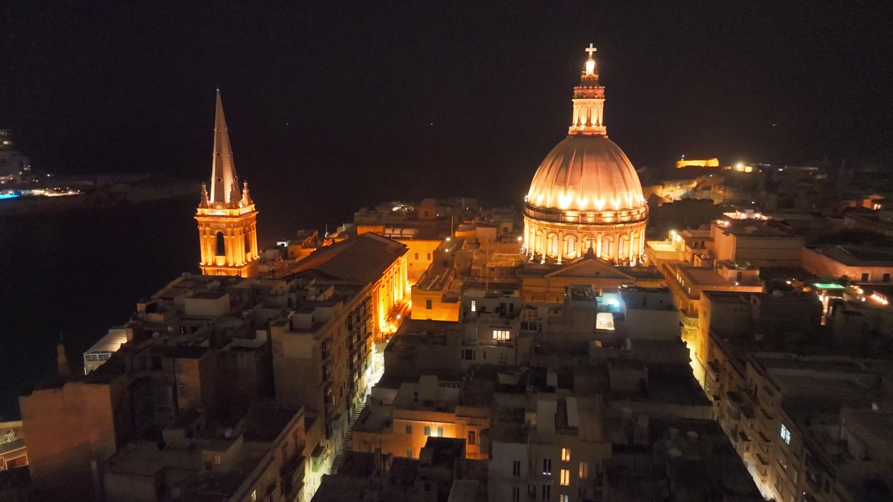 Aerial night view of Valletta Malta featuring narrow streets, illuminated dome and waterfront skyline with city lights reflecting on the harbor as drone moves through the historic urban area.