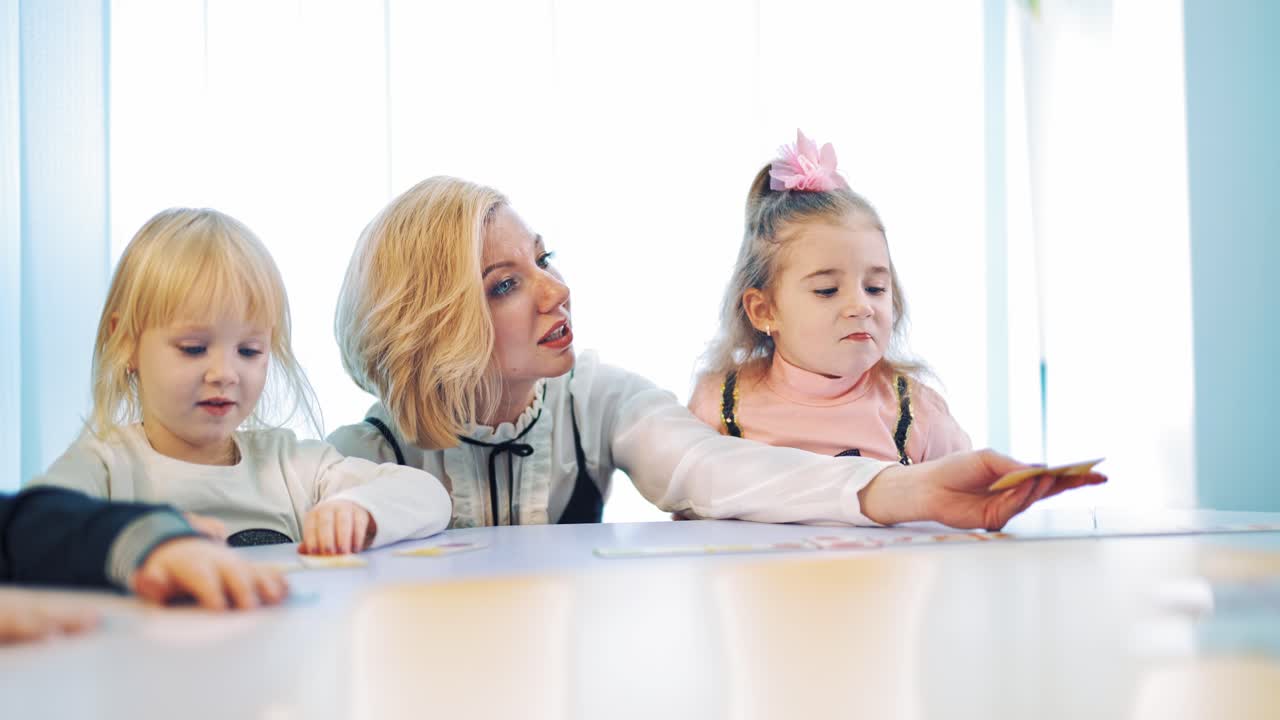 Little girls and their teacher at the table. Clever teacher talks to kids and develops their mind while working close to pupils. Woman plays with little children together at elementary school.