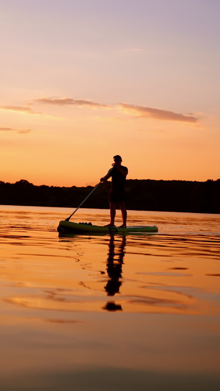 Dusk time on the calm river in summer. Man practices sup boarding alone in the nature. Low angle view. Vertical video.