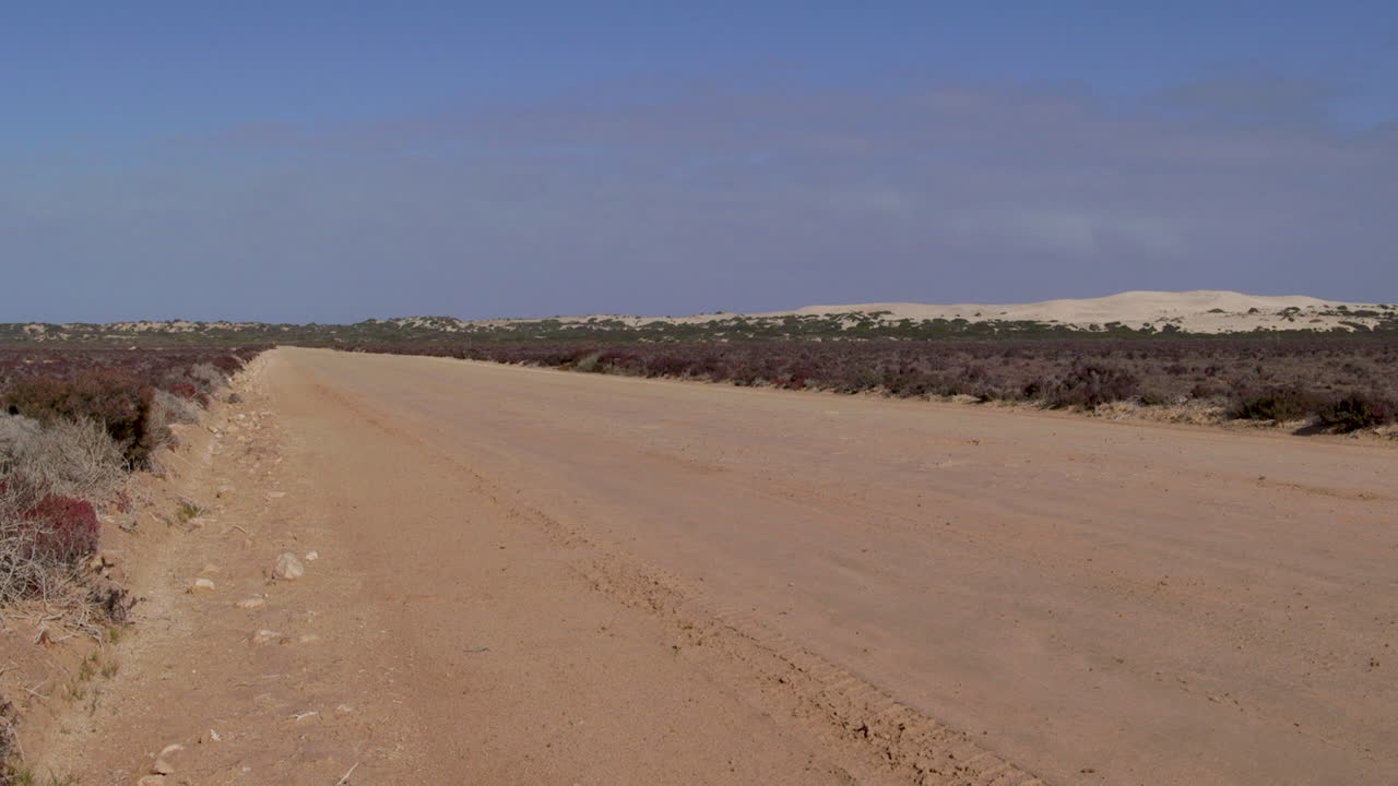 Uma longa e empoeirada estrada de cascalho estende-se em direção a dunas de areia distantes em uma paisagem árida.