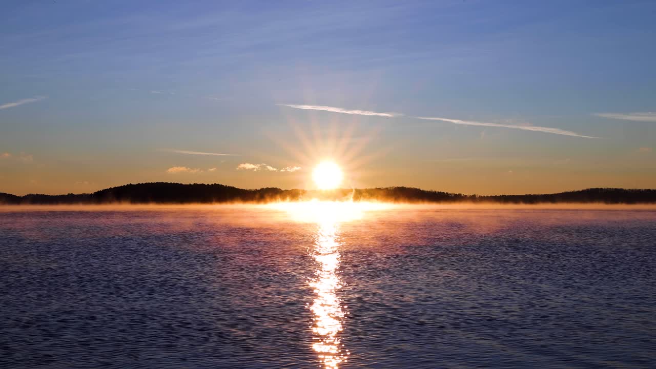 amanecer de 4k sobre un lago con montaña en el fondo, rayos de sol rociando a través del agua, cielo azul y naranja, niebla en el agua