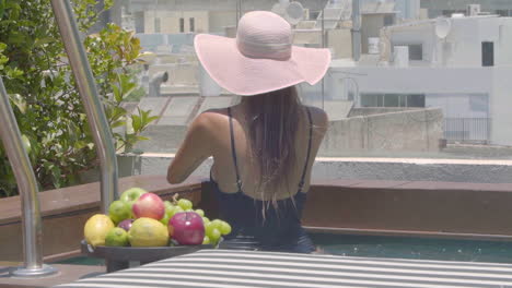 a woman in a black bathing suit and straw hat strolls by a private pool with a stunning city view