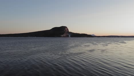 Flying-away-from-shore-towards-a-lone-mountain-across-a-lake-at-dawn-AERIAL