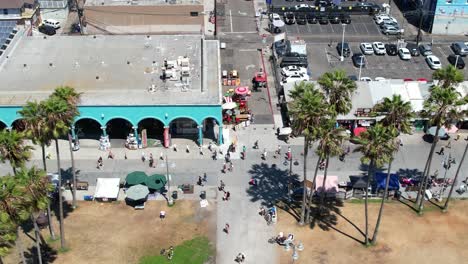 People-walking-along-the-famous-Venice-Beach-boardwalk-in-Los-Angeles,-California---sliding-aerial-flyover